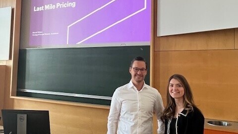 Rebecca B und Manuel W. stehen in einem Vorlesungsraum. Im Hintergrund ist die Präsentation des Vortrags zu erkennen