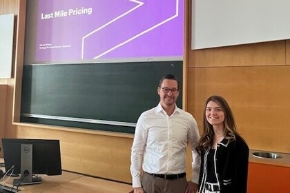 Rebecca B und Manuel W. stehen in einem Vorlesungsraum. Im Hintergrund ist die Präsentation des Vortrags zu erkennen