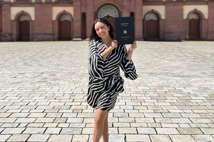 Yasmine Labib steht auf dem Ehrenhof und hält ihre Abschlussarbeit hoch. Sie hat lange dunkle Haare und trägt ein schwarz-weißes Kleid