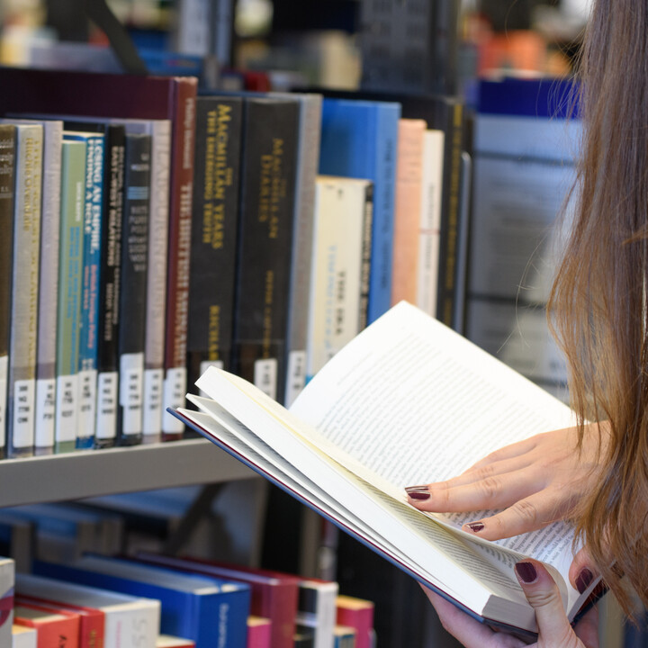 Eine Promovierende steht vor einem Bücherregal in der Bibliothek und liest in einem Buch
