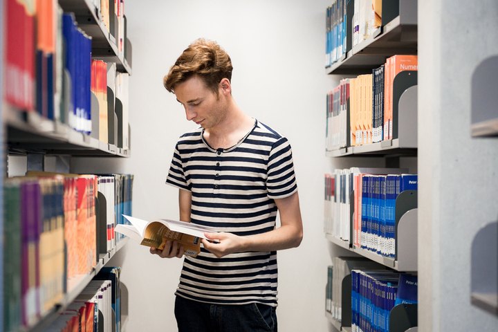 Eine Person mit kurzen Haaren und gestreiftem T-Shirt steht neben einem Bücherregal und liest in einem Buch