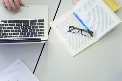 Ein Laptop, ein aufgeschlagenes Buch und eine Brille liegen auf dem Tisch.