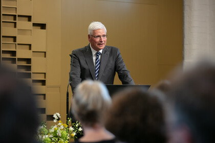 Eine Person mit kurzen grauen Haaren, einer Brille und einem grauen Anzug steht an einem Rednerpult cor Menschen, die verschwommen von hinten zu sehen sind