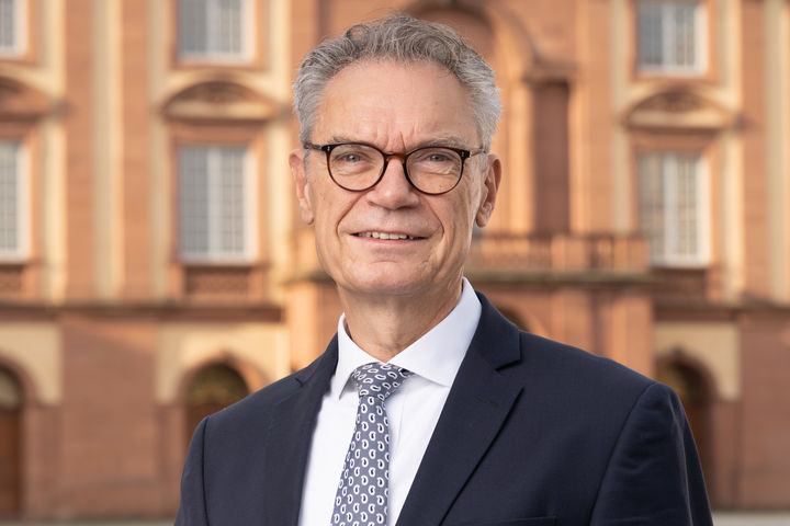 Joachim Lutz trägt einen Anzug und hat graue Haare und eine Brille. Hinter ihm ist das Schloss zu sehen