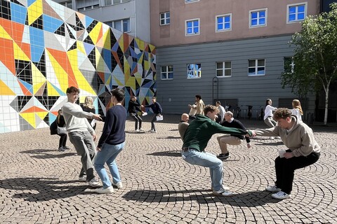 People in groups of two are holding hands and leaning backwards. In the background is a colorfully painted wall.