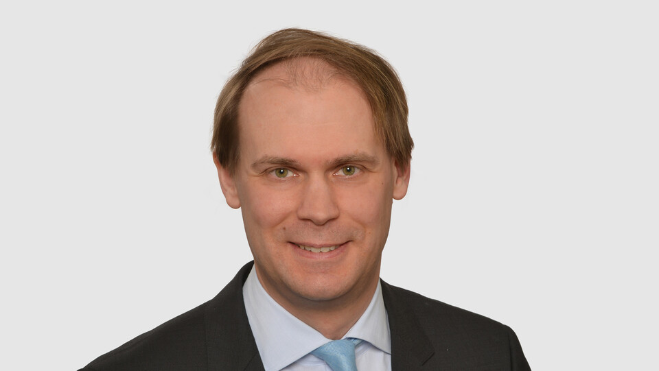 Prof. Bischof has short hair and wears a dark jacket, a light-colored shirt and a turquoise tie.