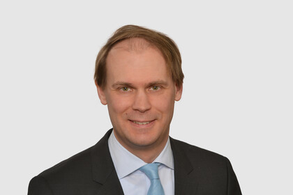 Prof. Bischof has short hair and wears a dark jacket, a light-colored shirt and a turquoise tie.