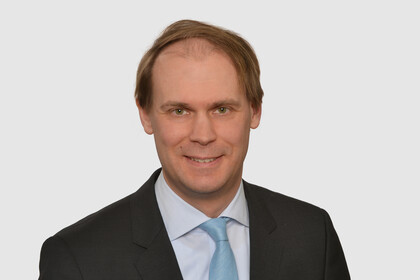 Prof. Bischof has short hair and wears a dark jacket, a light-colored shirt and a turquoise tie.