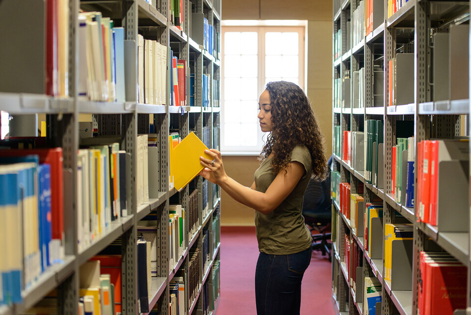 Eine Studentin zieht ein gelbes Buch aus einem Bücherregal in der Bibliothek.