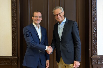 Dekan Lutz und Prof. Reinwald stehen vor einer dunklen Holztür und schütteln sich die Hände. Beide Lächeln in die Kamera.