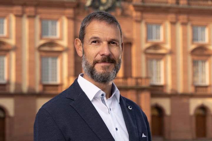 Ingo Bayer hat kurze Haare und einen Bart. Er trägt ein weißes Hemd und ein dunkles Sacco. Im Hintergrund ist das Schloss zu sehen.